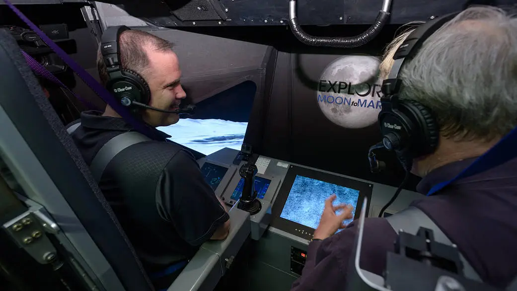 two men sitting inside simulation cockpit with a screen that says "explore moon to mars"